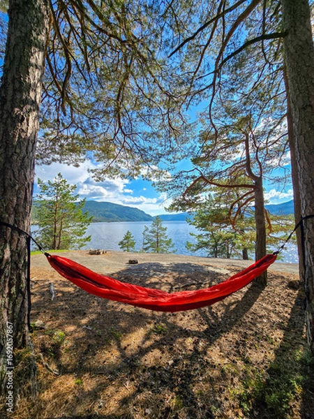 Fototapeta Vibrant red hammock sways gently between two tall trees, offering a perfect spot for relaxation. Lush greenery surrounds the tranquil waters of Norway, inviting peace and serenity.