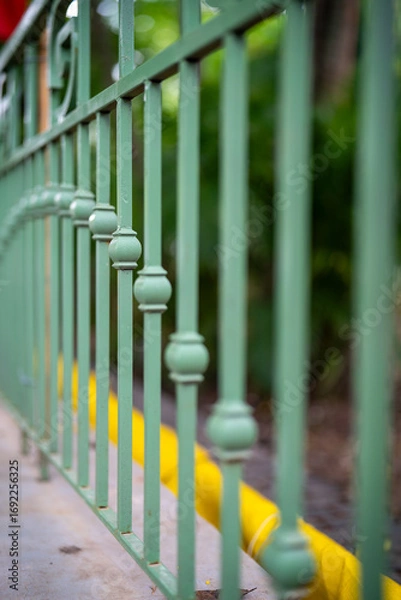 Fototapeta Close-up of green decorative railing with blurred background of plants and yellow curb.