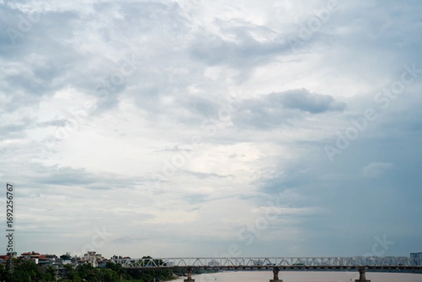 Fototapeta A serene view of a cloudy sky above a river with a bridge in the foreground.