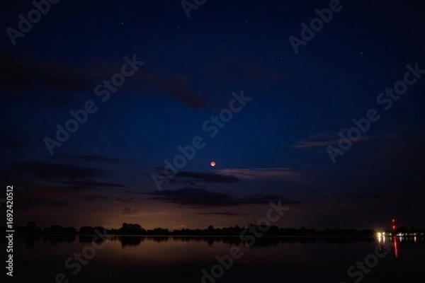 Fototapeta Blood moon shines over calm water reflecting distant lights under a dark blue night sky with clouds