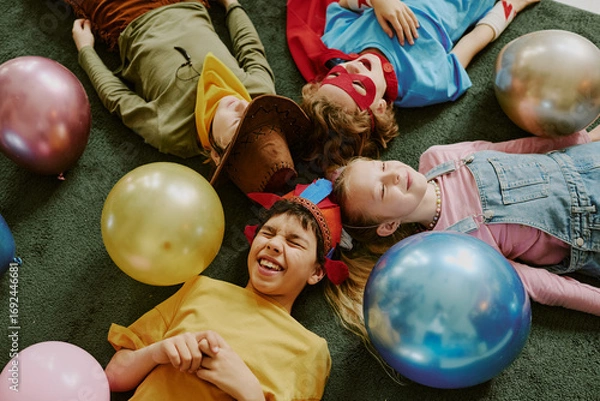 Fototapeta Group of four children lying on floor surrounded by balloons, two boys and two girls, diverse ethnicities, smiling and wearing costumes, celebrating together at party