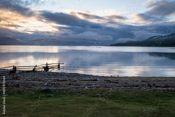 Fototapeta Dramatic sunset over the Beagle Channel in Tierra del Fuego, with reflections on the calm water, mountains in the background, and a rustic wooden fence on the shore