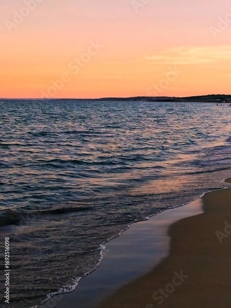 Fototapeta A serene view of a beach and sea at sunset or sunrise. The soft, warm colors of the sky (orange, pink, and peach) contrast delicately with the dark, cool tones of the water and wet sand.