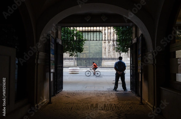 Fototapeta A man stands under an archway, looking out at a street with a blurred cyclist passing by, framed by intricate architecture in the distance.