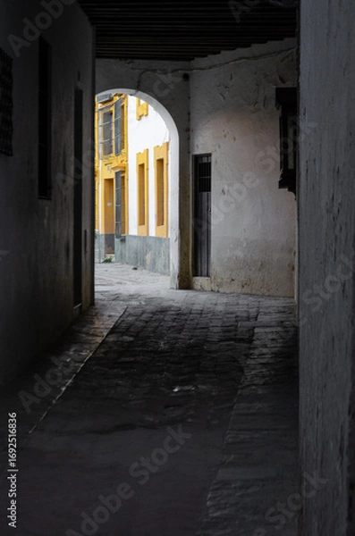 Fototapeta A quiet, shadowy alleyway opens up to a bright street in a historic town. The contrast highlights the hidden, secluded feel and the old architecture.
