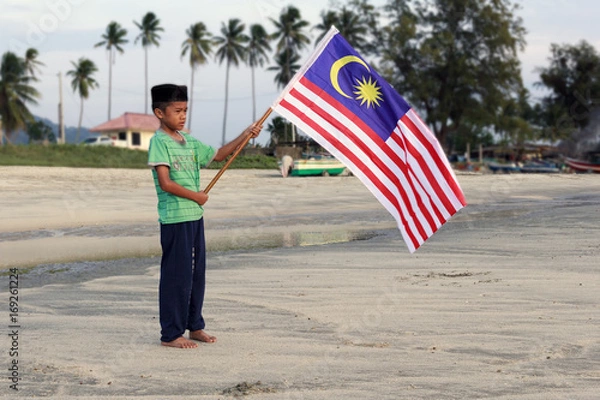Fototapeta The Concept of Independence Day - a boy holding the Malaysian flag on the shore