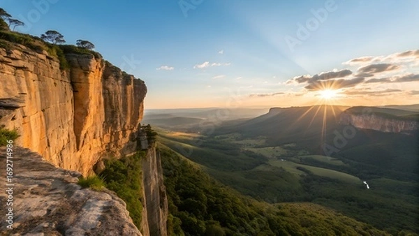 Fototapeta The golden sunset casts long rays over a magnificent valley, showcasing towering bluffs and lush green forests below a clear blue sky.