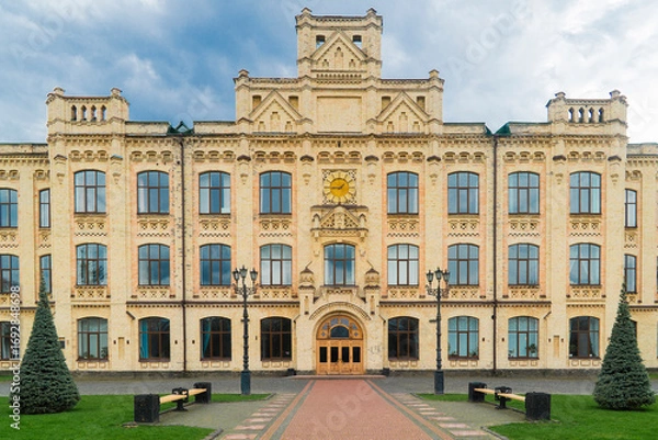 Fototapeta Kyiv. Kyiv Polytechnic Institute. Large, yellow brick building with detailed architectural features, numerous windows, and surrounding green lawns and trees on a cloudy day