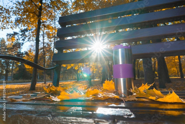 Fototapeta A stainless steel thermos mug with a purple insert stands on a park bench, surrounded by golden maple leaves with bright sun rays in the background