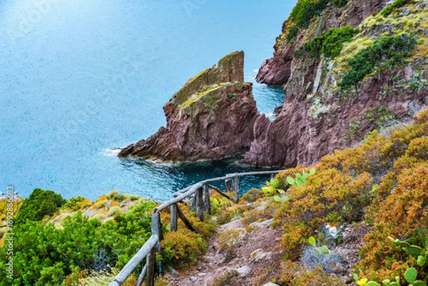 Fototapeta Breathtaking view of the turquoise Mediterranean Sea meeting the unique red rocks of Cala Zurletto in Capraia Island, with a wooden fence guiding the way along the cliffside vegetation