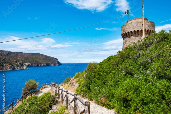 Fototapeta Wooden fence lining a gravel path leading to an ancient stone tower with the Italian flag waving, overlooking the blue sea and Capraia island's coastline on a sunny day