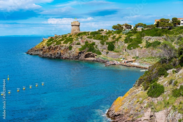 Fototapeta Turquoise waters washing rocky shores of Capraia Island, with a historic tower rising against a clear blue sky, create a tranquil and picturesque scene