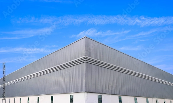 Fototapeta Warehouse factory industrial building with louver ventilation on gray aluminium corrugated metal and glass windows on white concrete wall against blue sky background, Low angle and symmetric view