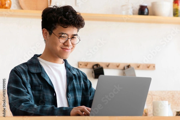 Fototapeta Modern remote job of blogger or employee and heath care during covid-19 lockdown. Cheerful asian young man in glasses works at laptop on table with coffee and water in boho style kitchen interior