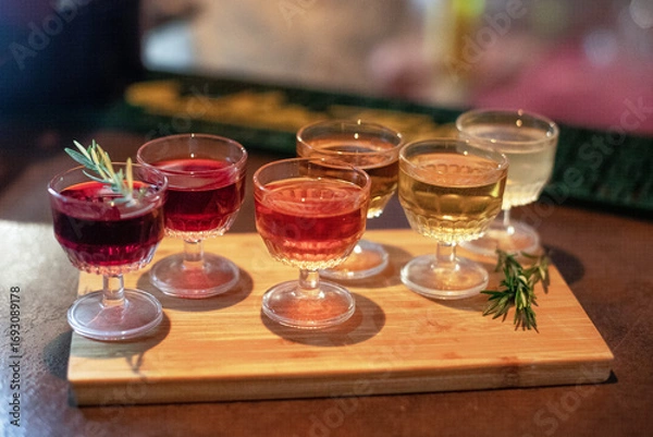Fototapeta A selection of colorful cocktails served in vintage glassware on a wooden tray, garnished with sprigs of rosemary, creating a cozy and inviting atmosphere