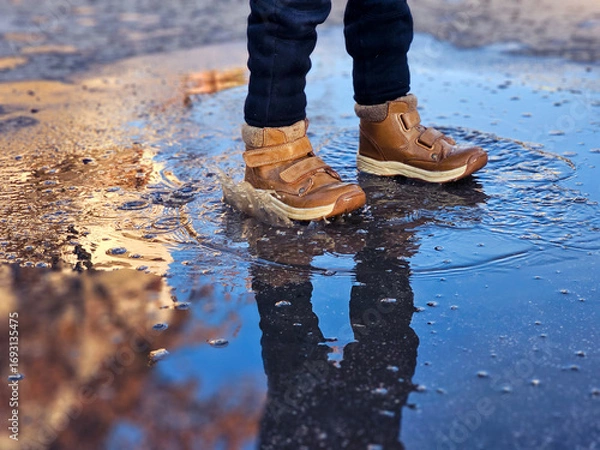 Fototapeta Child’s brown boots splashing in a puddle on a wet street. Close-up action shot capturing water droplets and playful movement outdoors.
