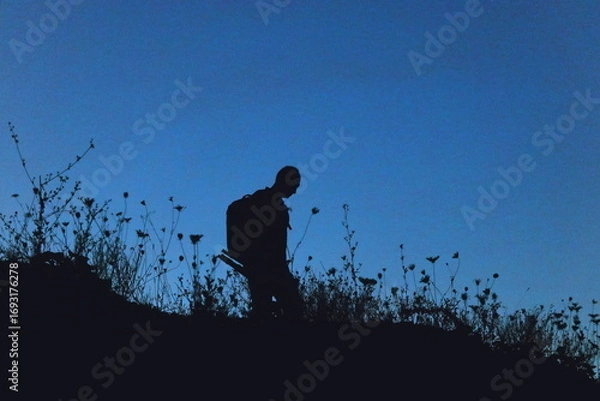 Fototapeta Silhouette of a lone hiker with a backpack standing on a hill at twilight, surrounded by wild grasses against a deep blue evening sky.