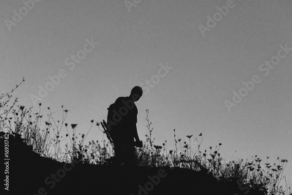 Fototapeta Silhouette of a lone hiker with a backpack standing on a hill at twilight, surrounded by wild grasses against a deep blue evening sky.