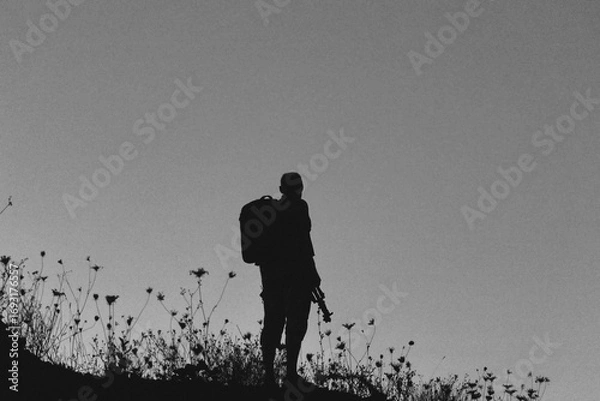 Fototapeta Silhouette of a lone hiker with a backpack standing on a hill at twilight, surrounded by wild grasses against a deep blue evening sky.