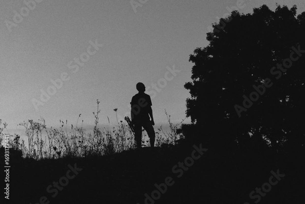 Fototapeta Silhouette of a lone hiker with a backpack standing on a hill at twilight, surrounded by wild grasses against a deep blue evening sky.