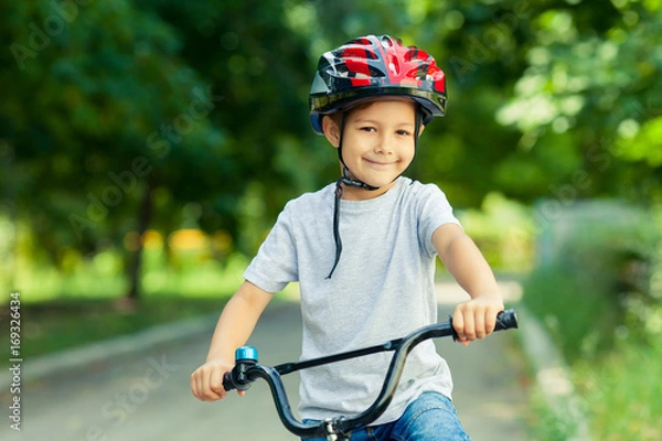 Fototapeta Little boy learns to ride a bike in the Park near the home. Portrait of a cute kid on bicycle. Happy smiling child in helmet riding a cycling.