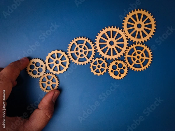 Fototapeta The image shows a hand placing a wooden gear, which is part of a larger arrangement of gears on a blue background