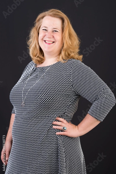 Obraz Portrait of a attractive european light overweighted red haired female in white black dress smiling friendly in front of black backgrouind - half body studio shot