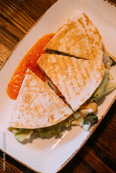 Fototapeta Tortilla food sliced. Served burrito. Grilled stuffed flatbread with lettuce. Hot meal cut piece with red spicy sauce on dish table.