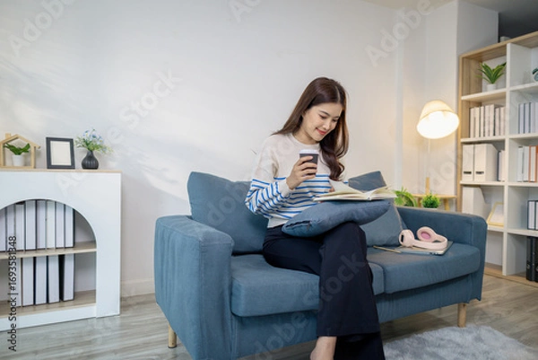 Fototapeta A young woman sits comfortably on a sofa, reading a notebook while holding a coffee cup. She looks relaxed and focused, enjoying a calm moment of study or planning at home.