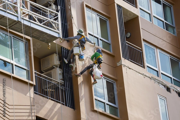 Fototapeta Two workers in safety gear perform maintenance on high rise building facade, using ropes and harnesses for support