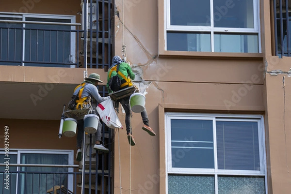 Fototapeta Workers repairing building facade with safety harnesses, using buckets and tools. Urban maintenance scene