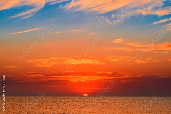Fototapeta The sun sinks into the sea surrounded by clouds, late evening over a warm sea, the splendor of autumn sunsets during the period of soft and warm velvet autumn