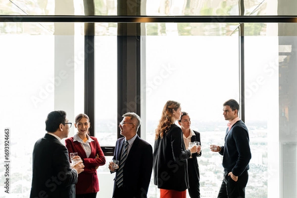Fototapeta Business professionals networking in a modern high-rise with panoramic city views, chatting with drinks during a corporate event by large windows.