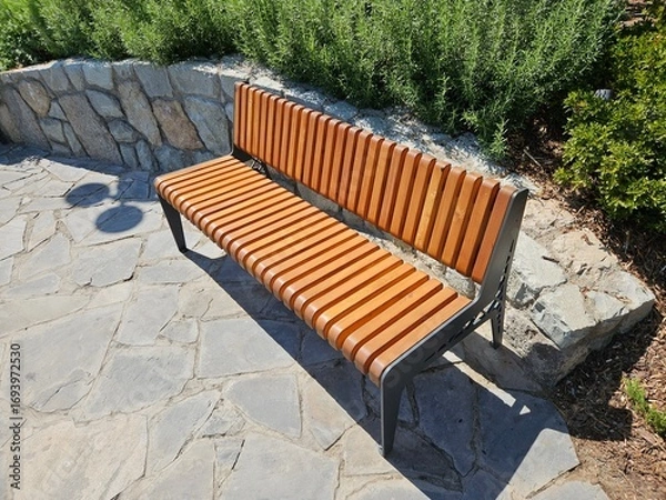 Fototapeta A modern wooden and metal bench sits in a sunny park on a stone patio. Ideal for urban planning, landscape architecture, or content about city recreation and public spaces.