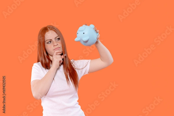 Fototapeta Woman contemplating finances while holding blue piggy bank against a bright orange background in a casual setting during the day