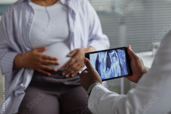 Fototapeta Pregnant Black woman sitting with hands on belly while medical professional holding tablet displaying ultrasound image during prenatal checkup in clinical setting