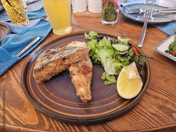 Fototapeta Crispy fried fish fillet with fresh salad and lemon on a rustic plate. Perfect for restaurant menus, food blogs, or healthy eating and pescatarian diet concepts.