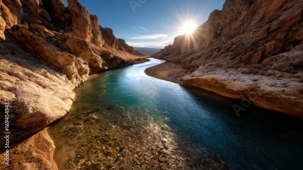 Fototapeta An enchanting view of a winding river flowing through a majestic canyon, beautifully illuminated by the golden light of the setting sun, highlighting the rugged textures of the cliffs.