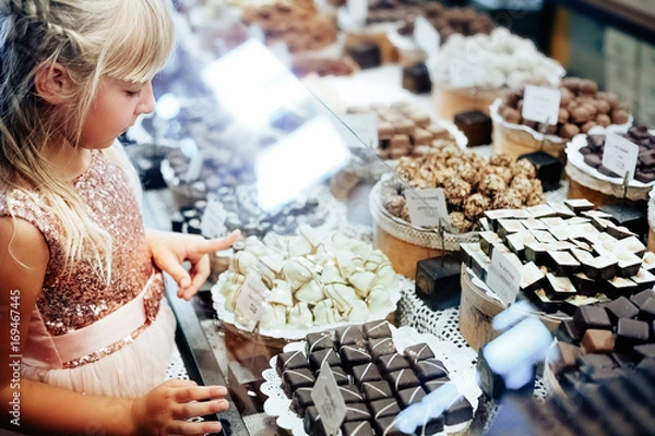 Fototapeta A little blonde girl is standing near a shop window with an assortment of sweets. The child chooses sweets