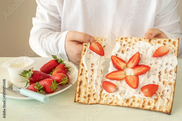 Obraz Matzah covered with creamy cheese and sliced strawberries in the hands of a child for tasty breakfast during Jewish holiday Pesach or Passover celebration.