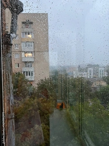 Obraz Rainy day city view through window glass with raindrops and blurred buildings creating moody atmosphere