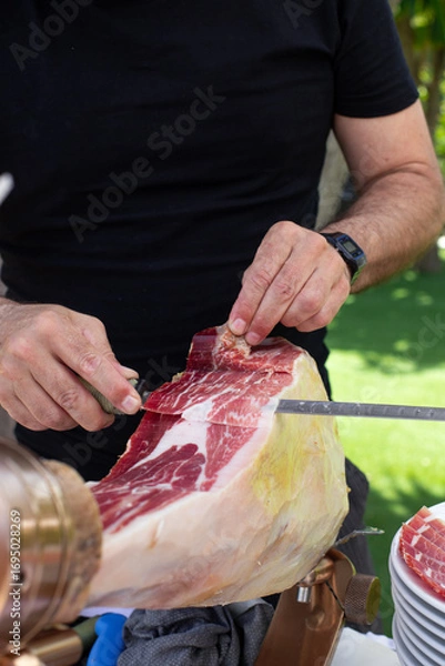 Obraz Man slicing traditional Iberian ham by hand with a long knife at an outdoor event
