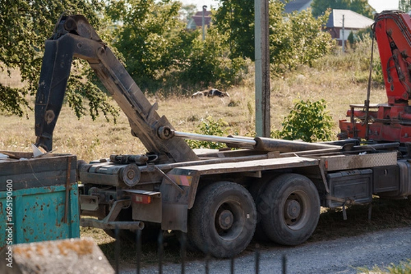Fototapeta Construction Vehicle with Crane Unloading Materials on Grassy Field, Red and Green Industrial Scene