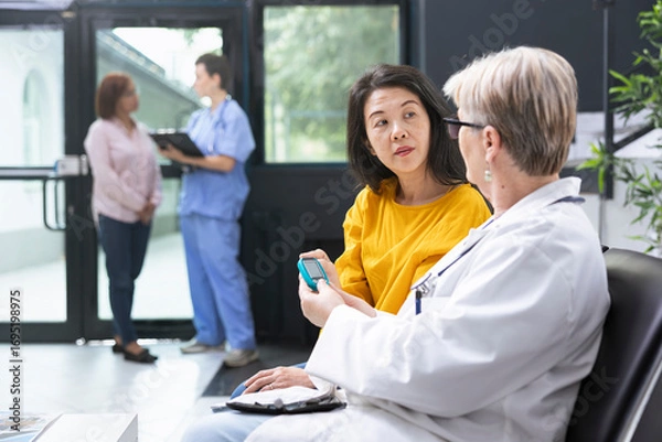Fototapeta Medical assistant conducting blood sugar levels measurement with glucometer during routine medical exam in clinic lobby. Asian patient discussing glucose readings and preventive care steps.