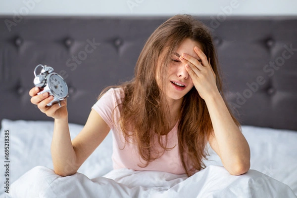 Fototapeta Depressed young woman with alarm clock sitting on bed, crying in despair, unwilling to get up to work, indoors. Exhausted millennial lady oversleeping, being late, feeling stressed and sleepy