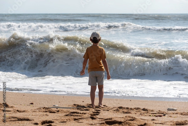 Obraz Caucasian child on a beach and watching big waves, fascinated by the ocean.