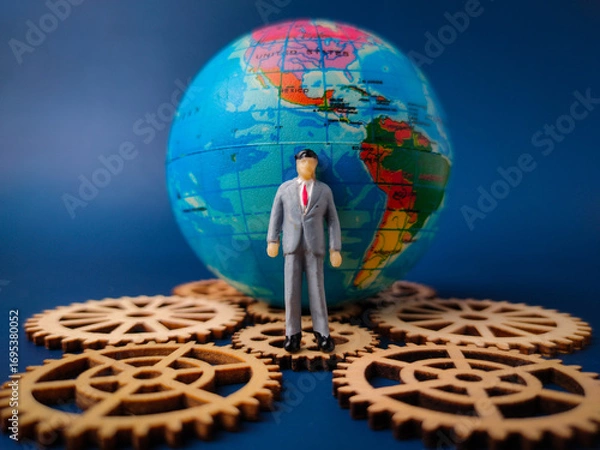 Fototapeta The image depicts a miniature businessman figure standing in front of a globe, with several wooden gears surrounding the base of the globe