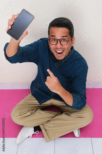 Fototapeta High angle view of asian man sitting cross legged holding blank phone display screen with happy face