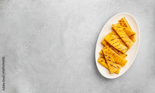 Fototapeta Grilled pineapple slices are arranged on a pristine white plate, top view, copy space