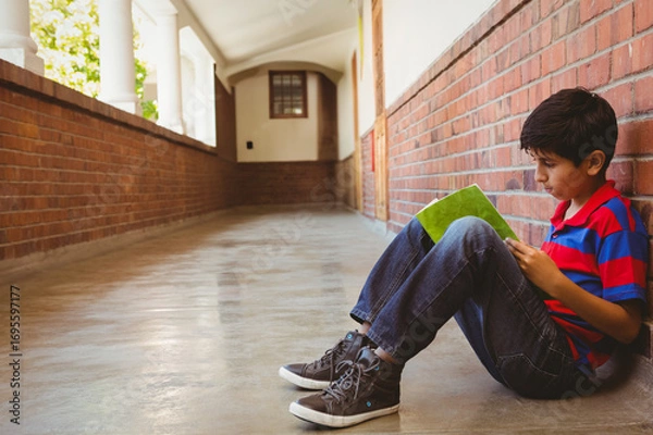 Obraz Boy sitting in brick corridor reading green book near white columns with greenery beyond
